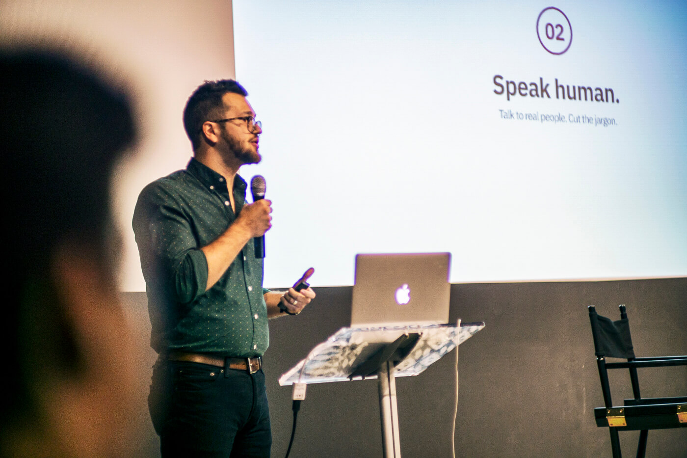 Jeremy speaks into a microphone presenting in front of a slide that reads 'Speak Human'.