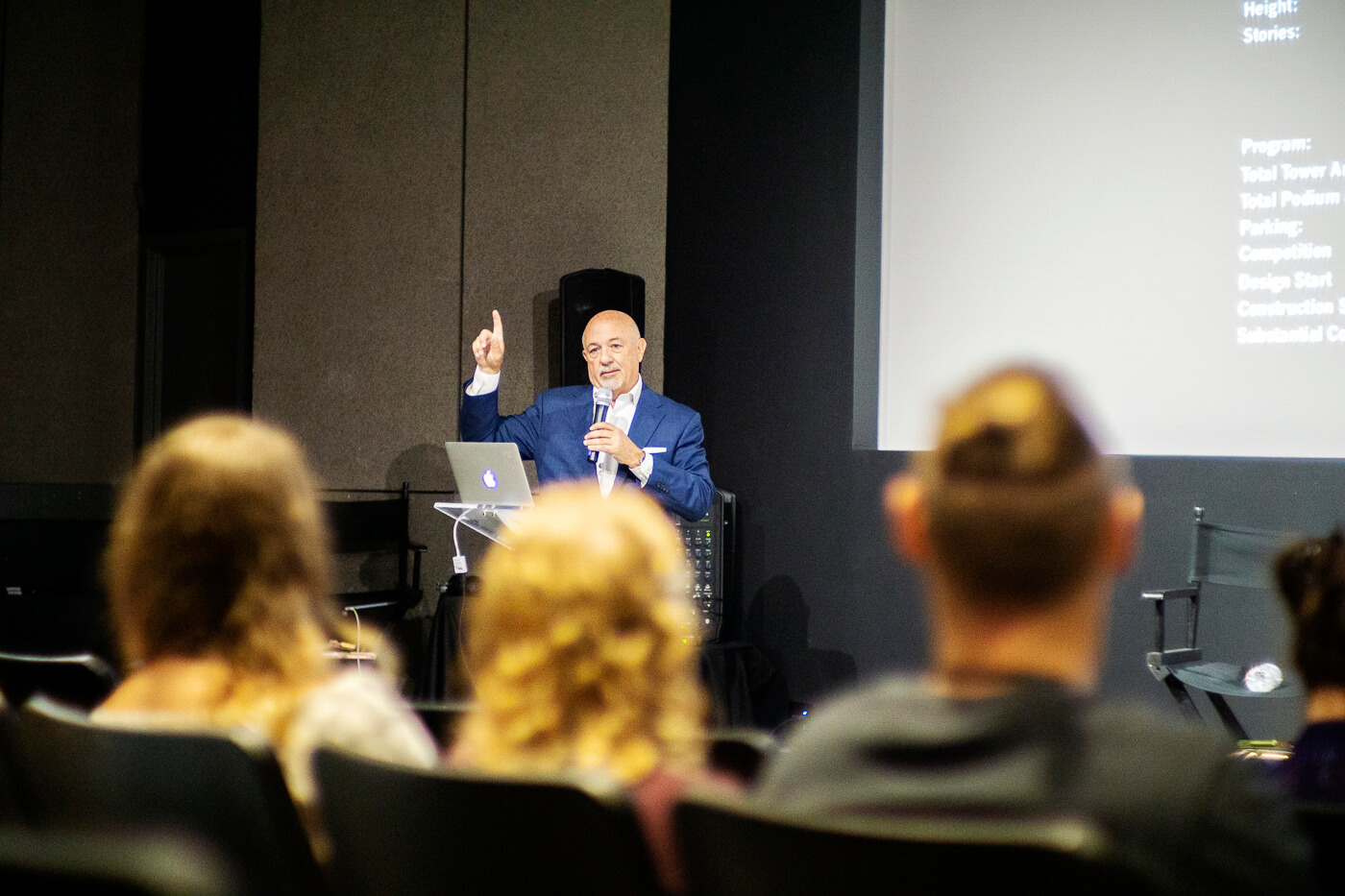 Viewed from the audience, George talks into a microphone and points upward while presenting.