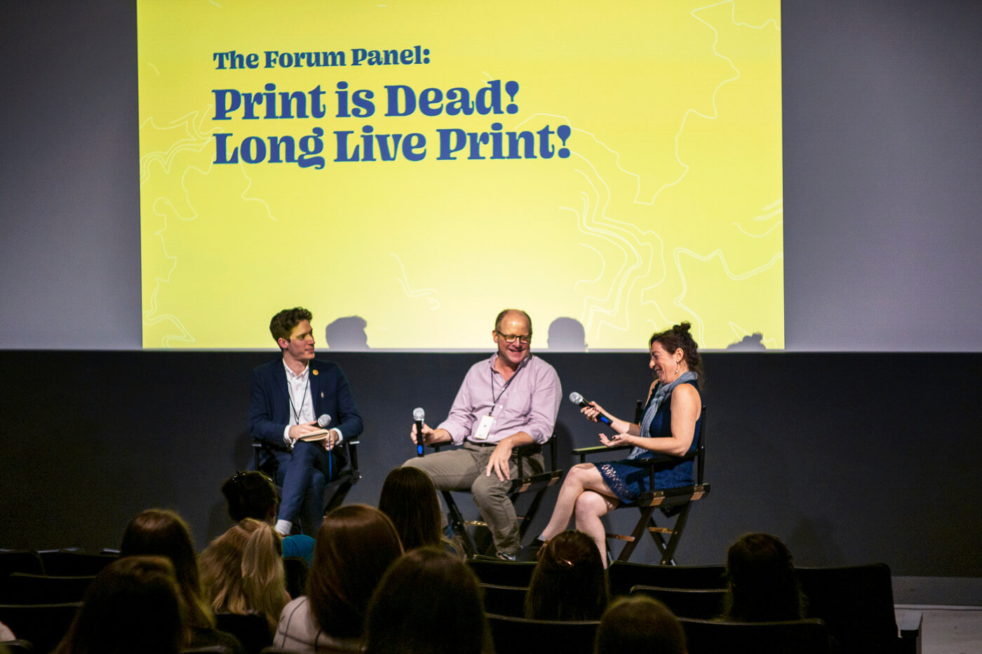Three people engaged in discussion sit in front of an audience and a screen reading 'The Forum Panel: Print is Dead! Long Live Print!'.