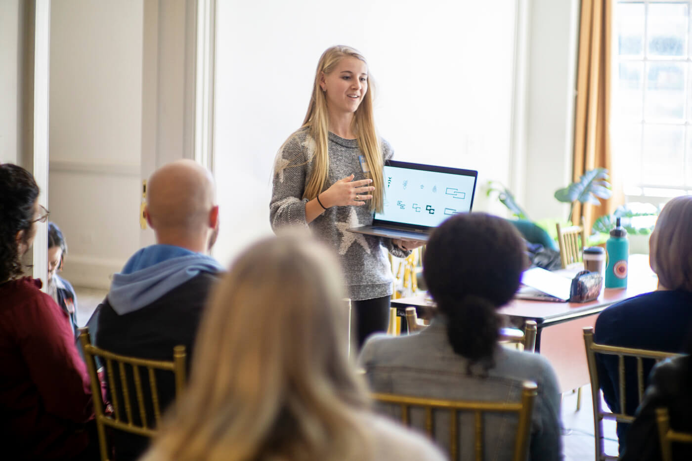 A girl holds a laptop and speaks in front of a small audience.